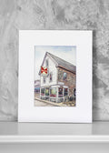 A matted watercolor art print depicting a quaint white and brown provincetown bat with a red and white sign, displayed against a textured gray wall.