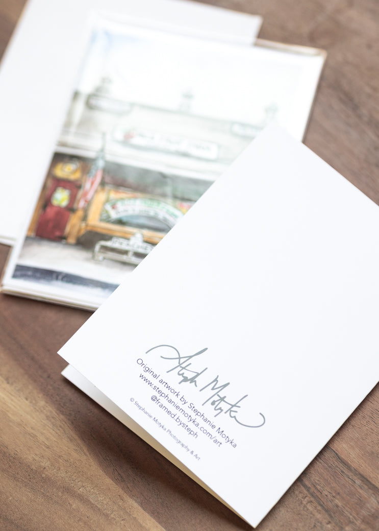 A white art print or postcard with a gray script signature and small printed text, placed on a wooden surface next to another printed card showing a street scene.