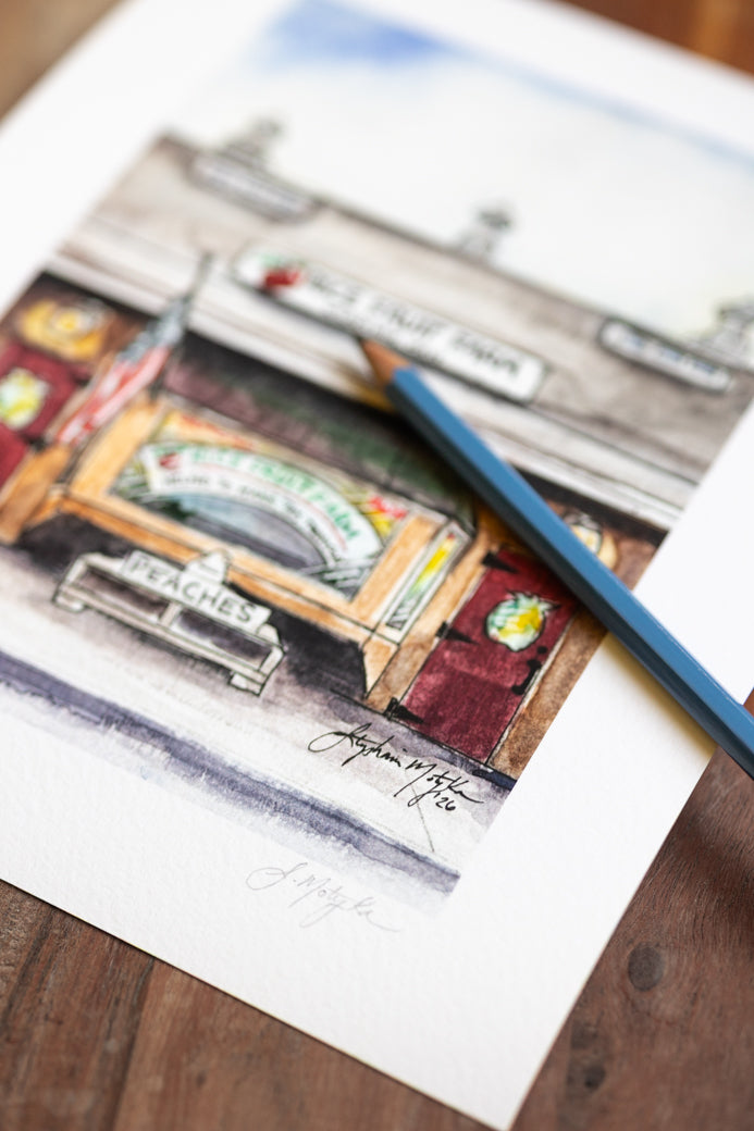 A watercolor art print depicting a vintage peaches market stall, shown on a white sheet of paper resting on a wooden surface with a blue pencil placed diagonally across it.