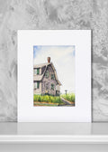 A matted art print featuring a watercolor painting of a weathered gray house with green grass and a blue sky, displayed against a textured gray wall.