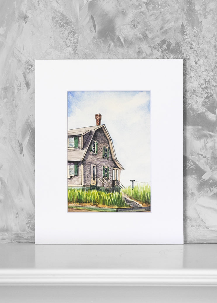 A matted art print featuring a watercolor painting of a weathered gray house with green grass and a blue sky, displayed against a textured gray wall.