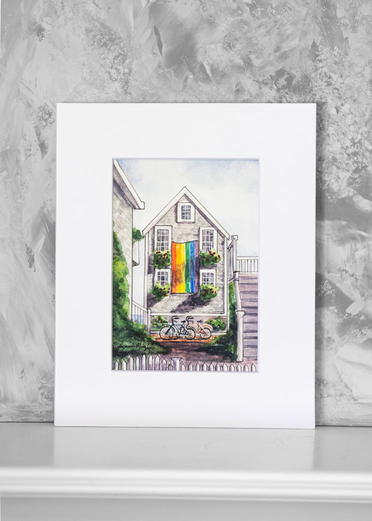 A matted art print featuring a watercolor painting of a house with a vibrant rainbow-colored flag displayed against a textured gray wall.