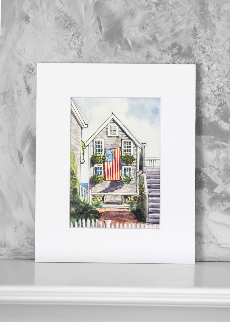 A matted watercolor art print depicting a coastal house with an American flag on the front, leaning against a textured gray wall on a white surface.