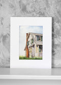 A watercolor art print of a gray house with a red brick chimney, displayed against a textured gray wall on a white surface.