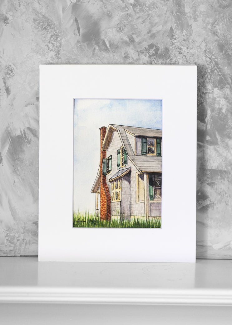A watercolor art print of a gray house with a red brick chimney, displayed against a textured gray wall on a white surface.
