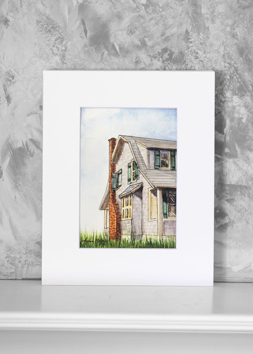 A watercolor art print of a gray house with a red brick chimney, displayed against a textured gray wall on a white surface.