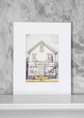 A matted art print featuring a watercolor illustration of a white bakery with storefront details, displayed against a textured gray wall on a white surface.