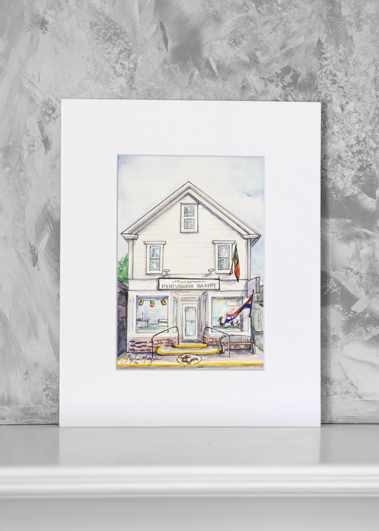 A matted art print featuring a watercolor illustration of a white bakery with storefront details, displayed against a textured gray wall on a white surface.