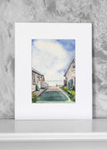 A watercolor art print depicting a coastal village walkway with houses and a blue sky, presented with a wide white mat board against a textured gray wall.