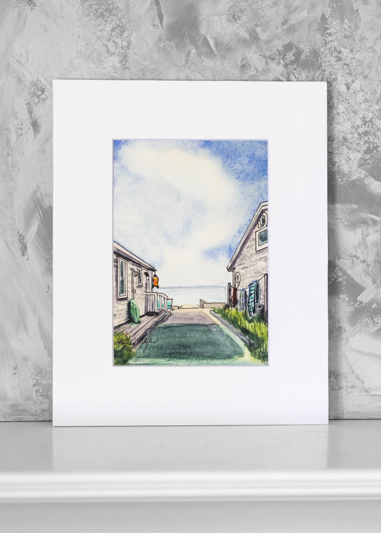 A watercolor art print depicting a coastal village walkway with houses and a blue sky, presented with a wide white mat board against a textured gray wall.