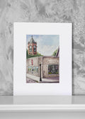 A matted art print featuring a watercolor painting of a brick building with a clock tower, displayed against a textured gray background.