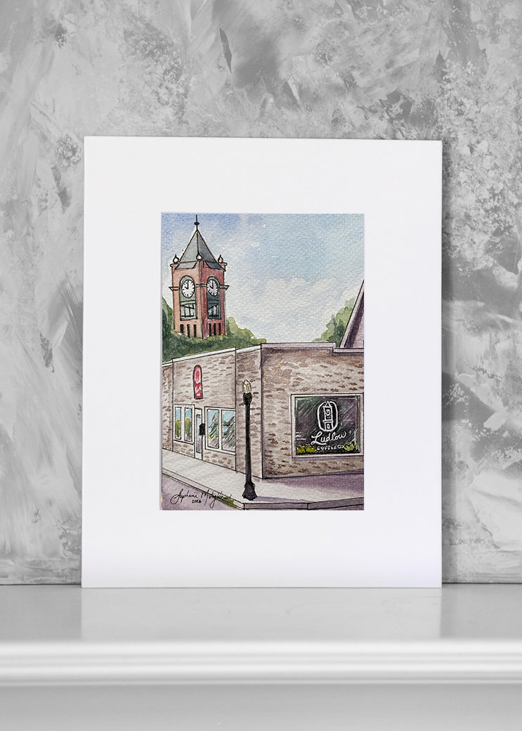 A matted art print featuring a watercolor painting of a brick building with a clock tower, displayed against a textured gray background.
