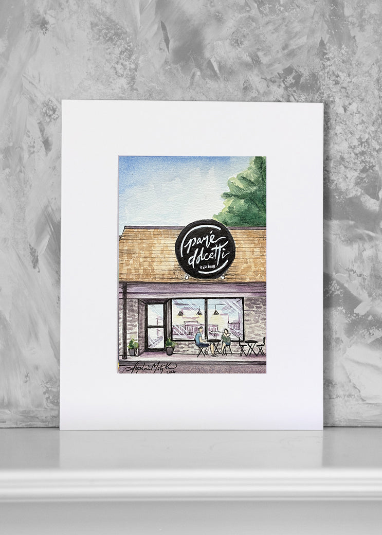 A matted art print featuring a watercolor illustration of a cafe storefront with the sign 'Pane Dolcetti, displayed against a textured gray wall.