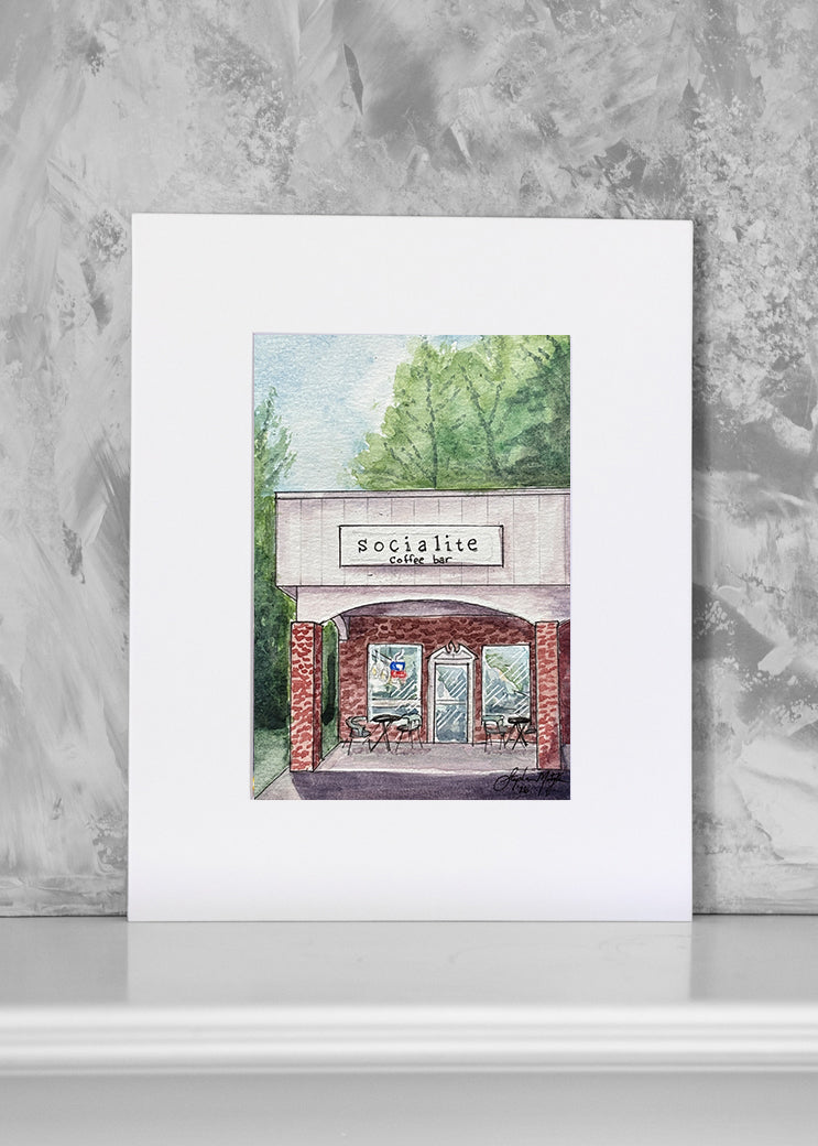 A matted art print featuring a watercolor illustration of a storefront with the sign 'socialite' above glass doors, set against a textured gray background.